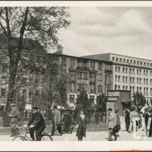 liczne osoby spacerujące lub przechodzące wzdłuż placu, umundurowany mężczyzna jedzie na rowerze, za nimi drzewa i pierzeja wypełniona wysokimi domami