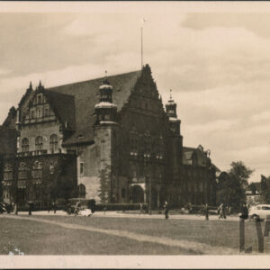 okazały gmach z wieloma szczytami i wieżami flankującymi wejście główne, na pierwszym planie trawnik i ulica, po której odbywa się ruch pieszych i pojazdów