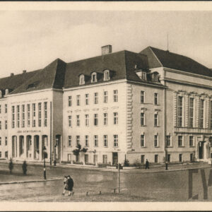 narożnik wielkiego, czterokondygnacyjnego gmachu o urozmaiconych elewacjach ze spadzistym dachem i wykuszem w fasadzie, przed nim fragment skrzyżowania ulic