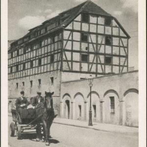 ulicą jedzie wóz konny powożony pzrez woźnicę, na wozie siedzi jeszcze jeden mężczyzna, w tle wysoki budynek z muru pruskiego i murowany płot