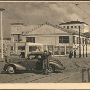 zabytkowy samochód stoi na pustej ulicy, obok niego policjant (?), w głębi hale targowe