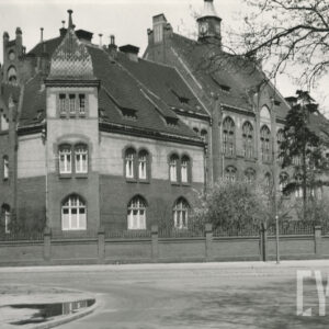 duży, licowany czerwoną cegłą, dwupiętrowy budynek, utrzymany w stylu neogotyckim, z dachem uwieńczonym wieżyczką, otoczony ceglanym płotem z metalowymi elementami