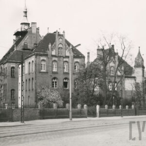 duży, licowany czerwoną cegłą, dwupiętrowy budynek, utrzymany w stylu neogotyckim, z dachem uwieńczonym wieżyczką i przybudówkami, otoczony ceglanym płotem z metalowymi elementami