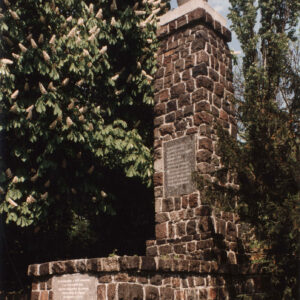 granitowy obelisk stojący na cokole, otoczony kamiennym murkiem i drzewami