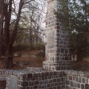 granitowy obelisk stojący na cokole, otoczony kamiennym murkiem i drzewami