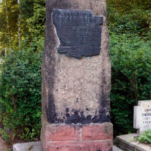 pomnik w formie steli o nieregularnych kształtach ustawionej na cokole z umieszczoną tablicą pamiątkową z napisem w języku polskim, obok groby i drzewa