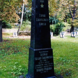 czarna tablica nagrobna z pozłacanym napisem cyrylicą w języku rosyjskim, wokół trawa, drzewa i inne nagrobki