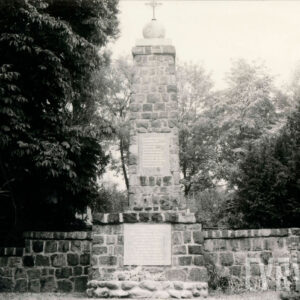 granitowy obelisk stojący na cokole, otoczony kamiennym murkiem i drzewami