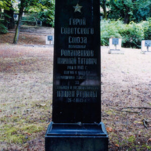 czarna tablica nagrobna z pozłacanym napisem cyrylicą w języku rosyjskim, wokół trawa, drzewa i inne nagrobki