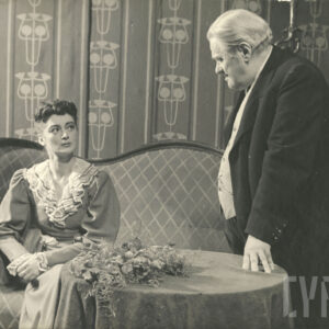 starszy, elegancko ubrany mężczyzna stojący przy stole obok elegancko ubranej kobiety siedzącej na kanapie