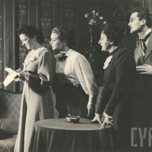 trzy elegancko ubrane kobiety, dwie młodsze, jedna starsza i jeden młody, elegancko ubrany mężczyzna; wszyscy stoją; jedna z kobiet czyta informację spisaną na kartce, pozostali stoją za nią i zaglądają jej przez ramię