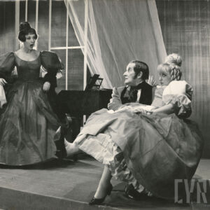 młoda kobieta siedząca na kolanach starszego mężczyzny, obok wzburzona druga kobieta