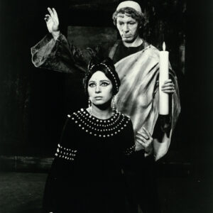 stojąca kobieta w mocnym makijażu, ubrana w suknię; za nią stoi mężczyzna w kostiumie nawiązującym do ubioru kapłana, unoszący jedną rękę w górę i trzymający w drugiej zapaloną świecę   