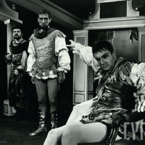 trzech mężczyzn w średnim wieku, jeden z nich siedzi na krześle i wskazuje palcem w kierunki dwóch pozostałych stojących obok