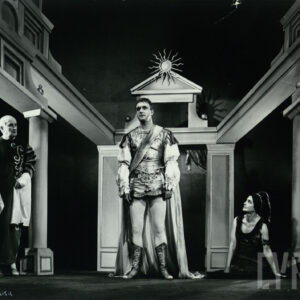 mężczyzna w średnim wieku w kostiumie nawiązującym do czasów bizantyjskich, po jego prawej stronie stoi inny, starszy mężczyzna, po lewej na ziemi siedzi kobieta