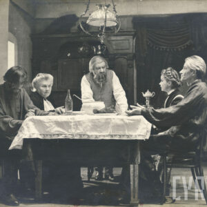 pięć osób przy nakrytym stole z jedzeniem, cztery z nich siedzą, jedna - starszy mężczyzna - stoi