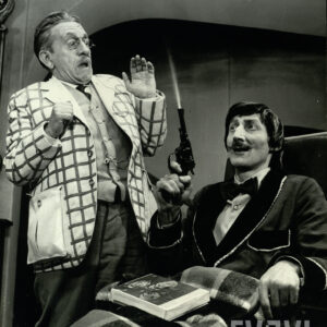 dwóch starszych, elegancko ubranych mężczyzn, jeden siedzi na wózku inwalidzkim z nogami przykrytymi kocem, w dłoni trzyma rewolwer; drugi mężczyzna stoi przerażony obok