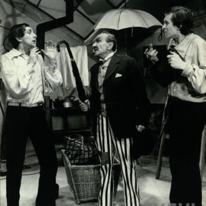 trzech elegancko ubranych mężczyzn stojących w rzędzie, mężczyzna w środku wygraża parasolem mężczyźnie stojącemu obok