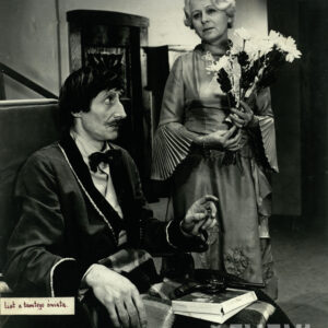 elegancko ubrany mężczyzna siedzący na wózku inwalidzkim z rewolwerem na kolanach, obok stoi starsza, elegancko ubrana kobieta z kwiatami w rękach