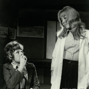 dwie kobiety w średnim wieku, jedna siedzi przy biurku, drga stoi obok