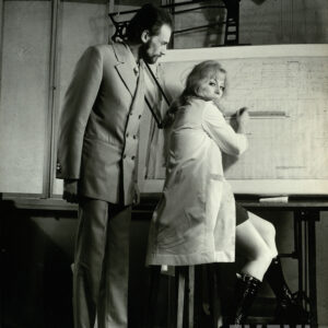 mężczyzna w średnim wieku ubrany w garnitur stojący obok kobiety siedzącej przy tablicy kreślarskiej; kobieta trzyma w rękach linijkę i przyrząd do pisania