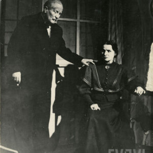 starszy mężczyzna w garniturze pochylający się nad siedzącą starszą kobietą ubraną w suknie