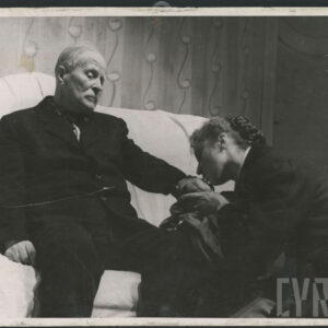 starszy mężczyzna w garniturze, siedzący na kanapie, przed nim klęczy kobieta całująca jego dłoń