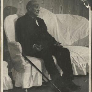 starszy mężczyzna w garniturze siedzący na kanapie, obok jego nóg leży laska