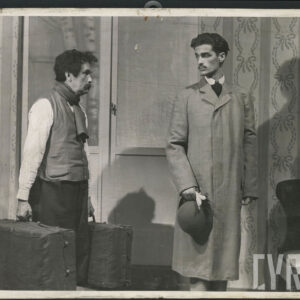 dwóch mężczyzn, jeden starszy, drugi młody spoglądających na siebie; starszy niesie walizki za elegancko ubranym młodym mężczyzna, który trzyma w rękach kapelusz