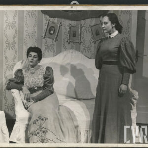 dwie kobiety w średnim wieku; jedna z nich, ubrana w elegancką suknię siedzi na kanapie; druga, ubrana w mniej odświętną suknię, stoi obok