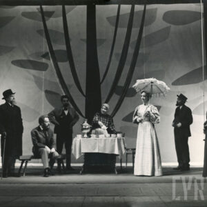 grupa odświętnie ubranych osób stojących przy stoliku z filiżankami i samowarem 