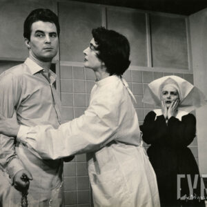 kobieta ubrana w fartuch lekarski, trzymająca mężczyznę za ramię, za nimi stoi zafrasowana kobieta w stroju siostry posługującej jako pielęgniarka w szpitalu