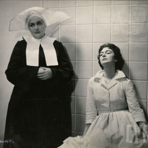 dwie kobiety w średnim wieku; jedna ubrana w fartuch lekarski, siedzi oparta o ścianę; obok stoi kobieta w stroju siostry posługującej jako pielęgniarka w szpitalu