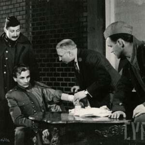 czterech mężczyzn, trzech z nich w mundurach wojskowych, jeden starszy w garniturze; jeden żołnierz siedzi przy stole, pozostali stoją 