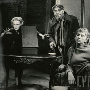 młoda kobieta siedzi na krześle, a za nią przy stole stoją starszy mężczyzna i kobieta w średnim wieku