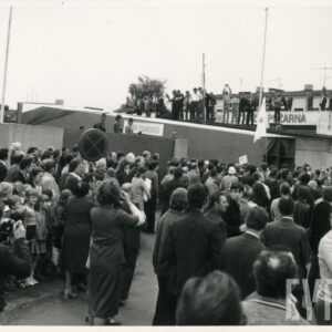 bardzo duża grupa ludzi czeka przed bramą zakładów, przez którą wyjeżdża duży element konstrukcyjny pomnika w formie zwężającego się prostopadłościanu