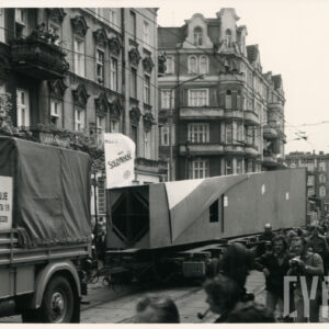 duży fragment pomnika w kształcie zwężającego się prostopadłościanu transportowany na platformie, wokół tłum ludzi, w głębi kamienice