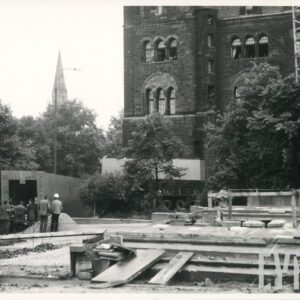 na pierwszym planie teren budowy, porozkładane deski, z lewej leżący na ziemi fragment pomnika, przy nim grupa mężczyzn, w głębi fragment dużego kamiennego budynki