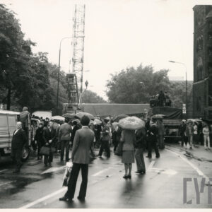 grupa ludzi, część pod parasolami, obserwuje mocowanie lin dźwigu do elementu konstrukcyjnego pomnika leżącego na platformie transportującej