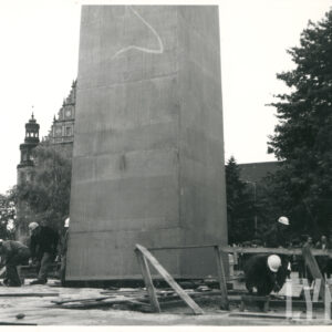 duży prostopadłościan, zwężający się ku górze usytuowany na przygotowanym pod niego fragmencie placu, wokół niego mężczyźni w białych kaskach