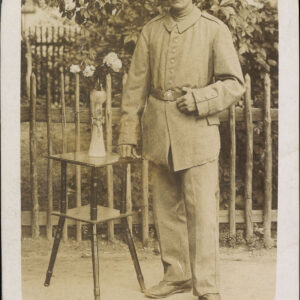 żołnierz w mundurze; po lewej stolik z kwiatami w wazonie; za drewnianym płotem drzewo