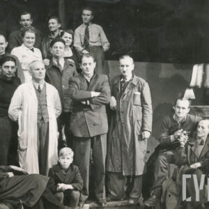 trzynaście osób pozujących na stojąco; z przodu pięć osób na siedząco; z po prawej stolik i lampa 