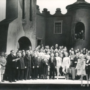 tłum ludzi na scenie; z tyłu scenografia zamku; mężczyzna po lewej czyta z kartki; pośrodku sceny mały stolik