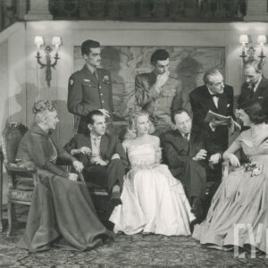 trzy kobiety i dwaj mężczyźni w eleganckich strojach siedzący na fotelach; z tyłu dwóch mężczyzn w mundurach; z prawej trójka mężczyzn pochylonych nad zeszytem