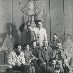 na siedząco sześciu mężczyzn i jedna kobieta; z tyłu stoi jeden mężczyzna; w tle scenografia teatralna