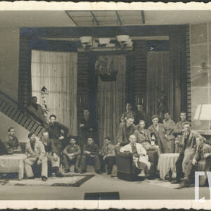 większa grupa ludzi w dużym pomieszczeniu; część z nich na siedząco na schodach i na fotelach ustawionych przy dwóch stołach, część na stojąco; po lewej schody do góry; z tyłu trzy wysokie okno; przy środkowym oknie model żaglowca