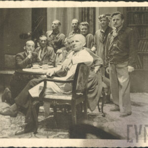 pięciu mężczyzn siedzących przy stole; za nimi na stojąco czterech mężczyzn i jedna kobieta; po prawej regał z książkami