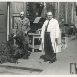 dwóch mężczyzn stojących przed ceglanym budynkiem; po lewej drzwi wejściowe; wzdłuż ściany ustawione części scenografii teatralnej