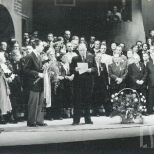 tłum ludzi na scenie; w tle scenografia; pośrodku mężczyzna czytający z kartki; na środku sceny kwiaty w koszu; po lewej mężczyzna z kwiatami