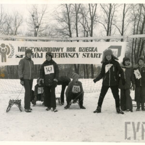 zimowa sceneria, grupa dzieci w zimowych ubraniach stoi na tle bramki do gry w piłkę nożną, kilkoro dzieci ma sanki